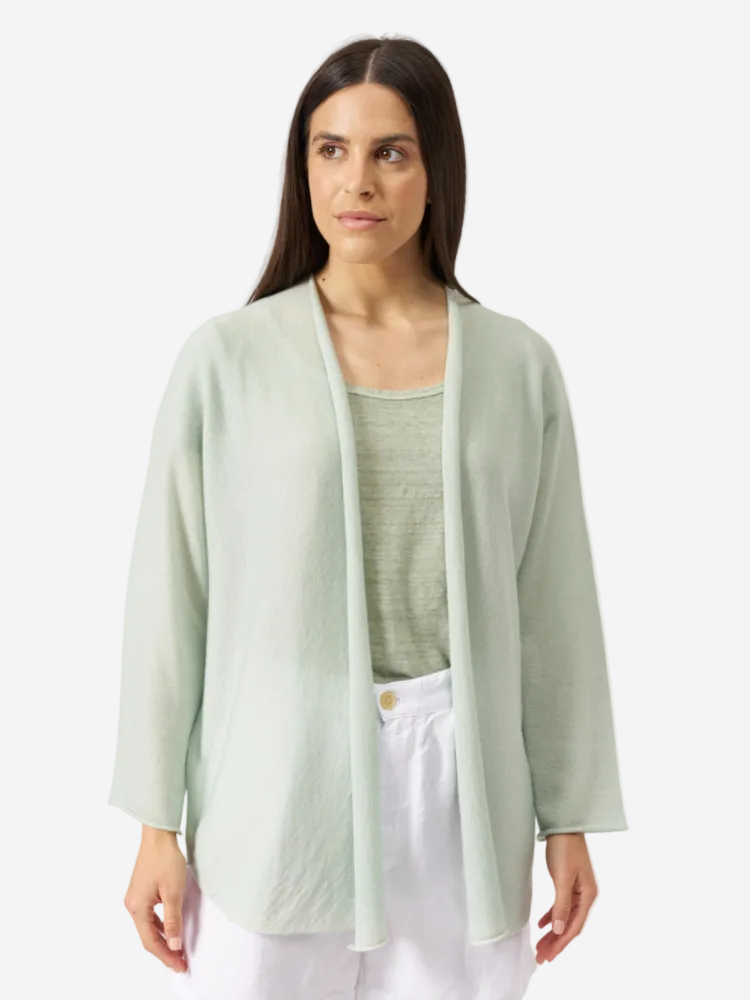 A woman wearing a green cashmere knitted cardigan with long sleeves, hip length, over a top and paired with white pants.