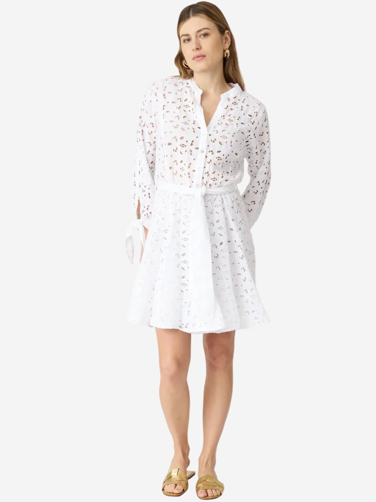 A woman standing and wearing a white mini dress with long sleeves and a V-neckline, paired with gold flat sandals.