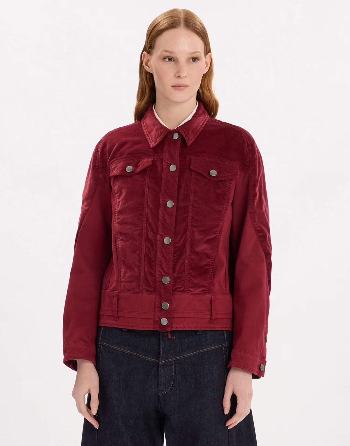 A model wearing a cropped, deep burgundy corduroy jacket with silver buttons, paired with dark blue jeans, against a plain white studio background.
