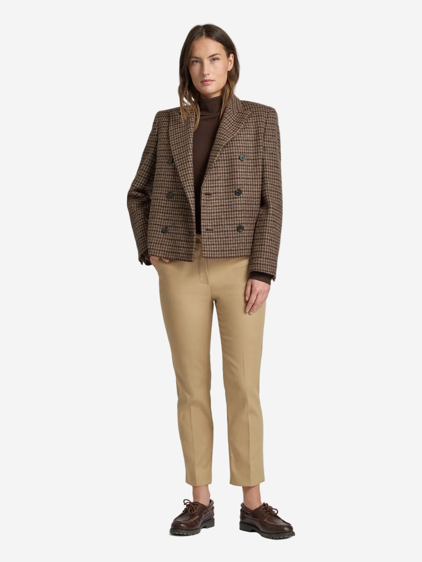 Woman wearing a brown checkered blazer, brown turtleneck, beige pants, and brown shoes on a light gray background