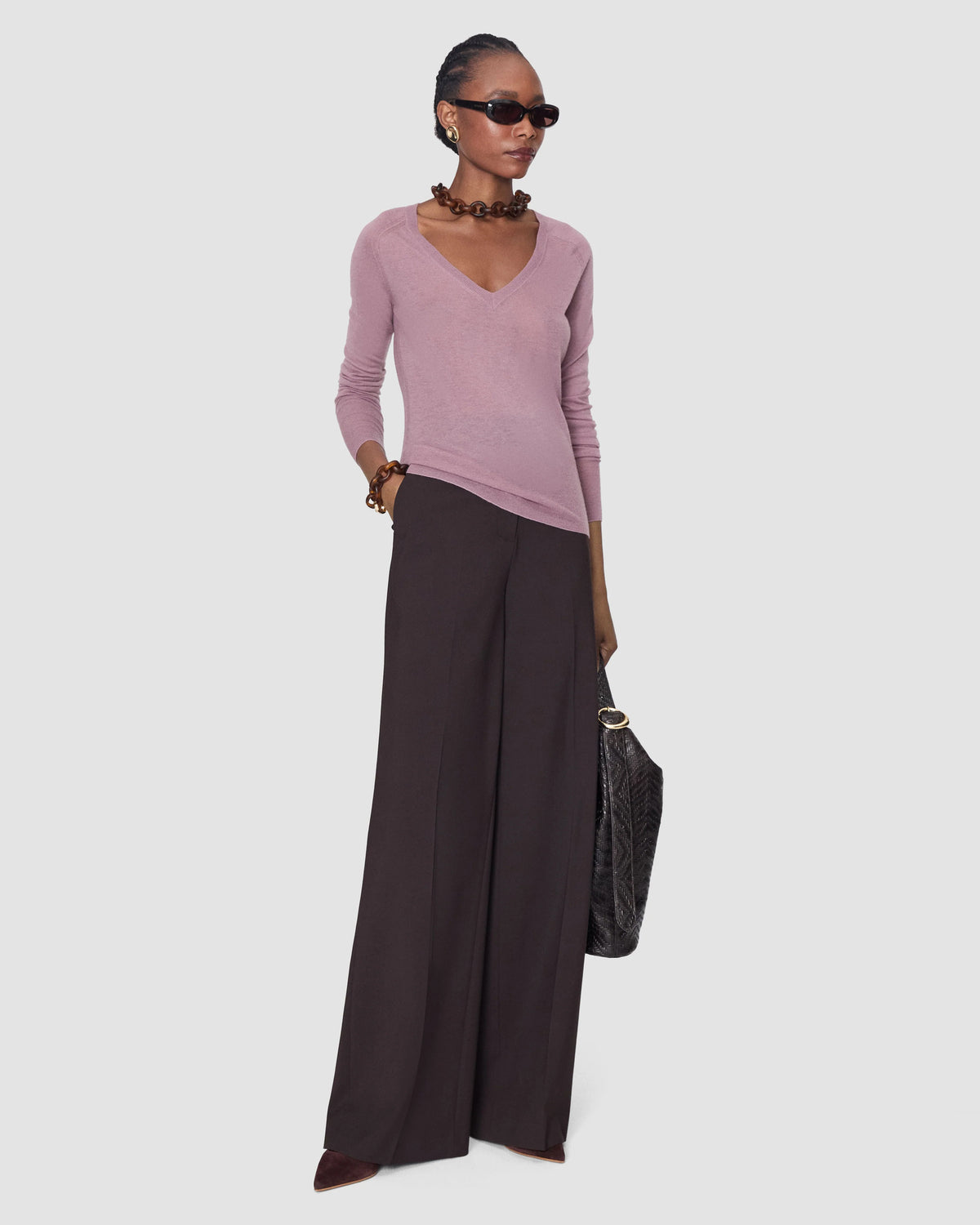 A model wearing a dusty pink long-sleeve V-neck knit top from Joseph and dark brown wide-leg trousers, accessorized with a chunky brown beaded necklace, sunglasses, bracelets, and a black textured handbag.