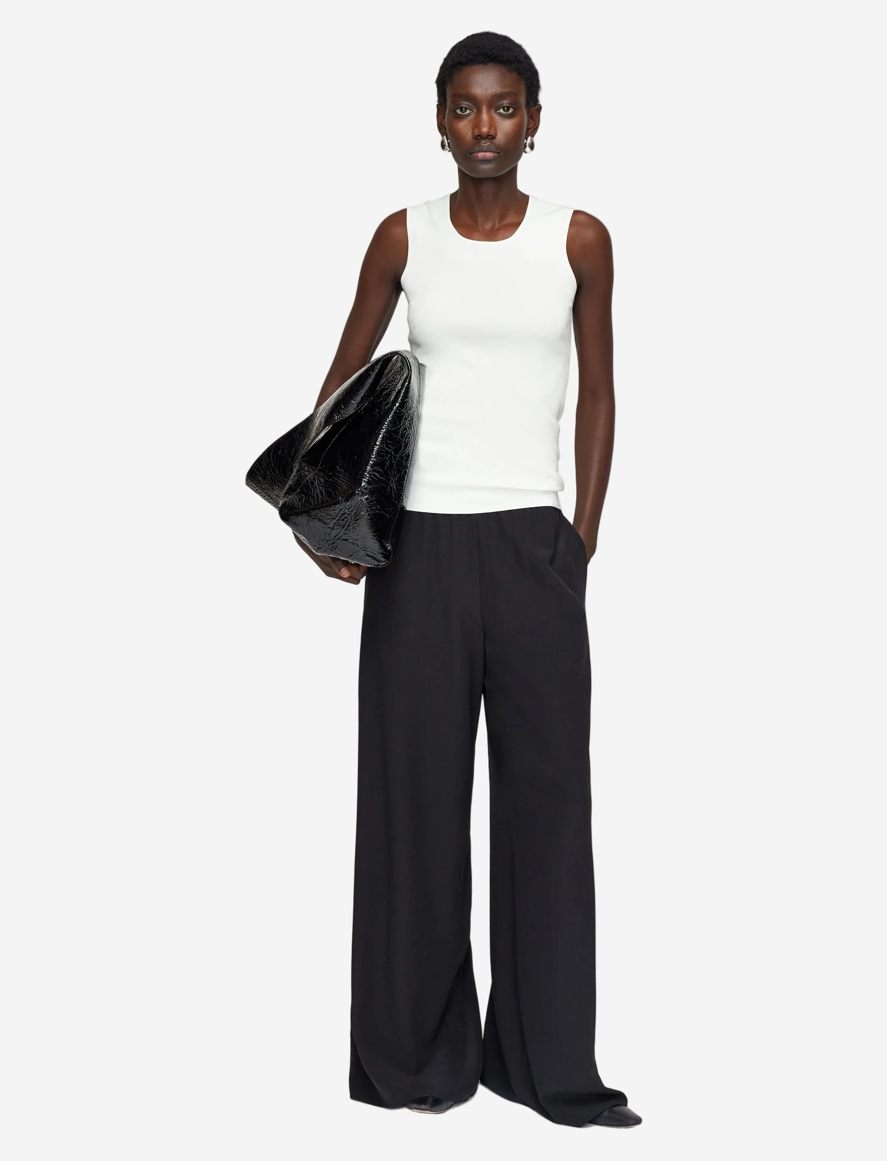 Woman wearing a white sleeveless top and black wide-leg pants holding a gray clutch against a light gray background