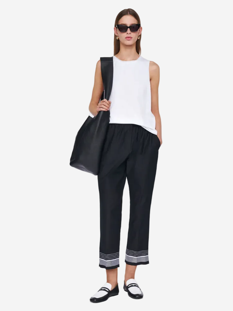 Woman wearing a white sleeveless top and black pants holding a black bag on a white background