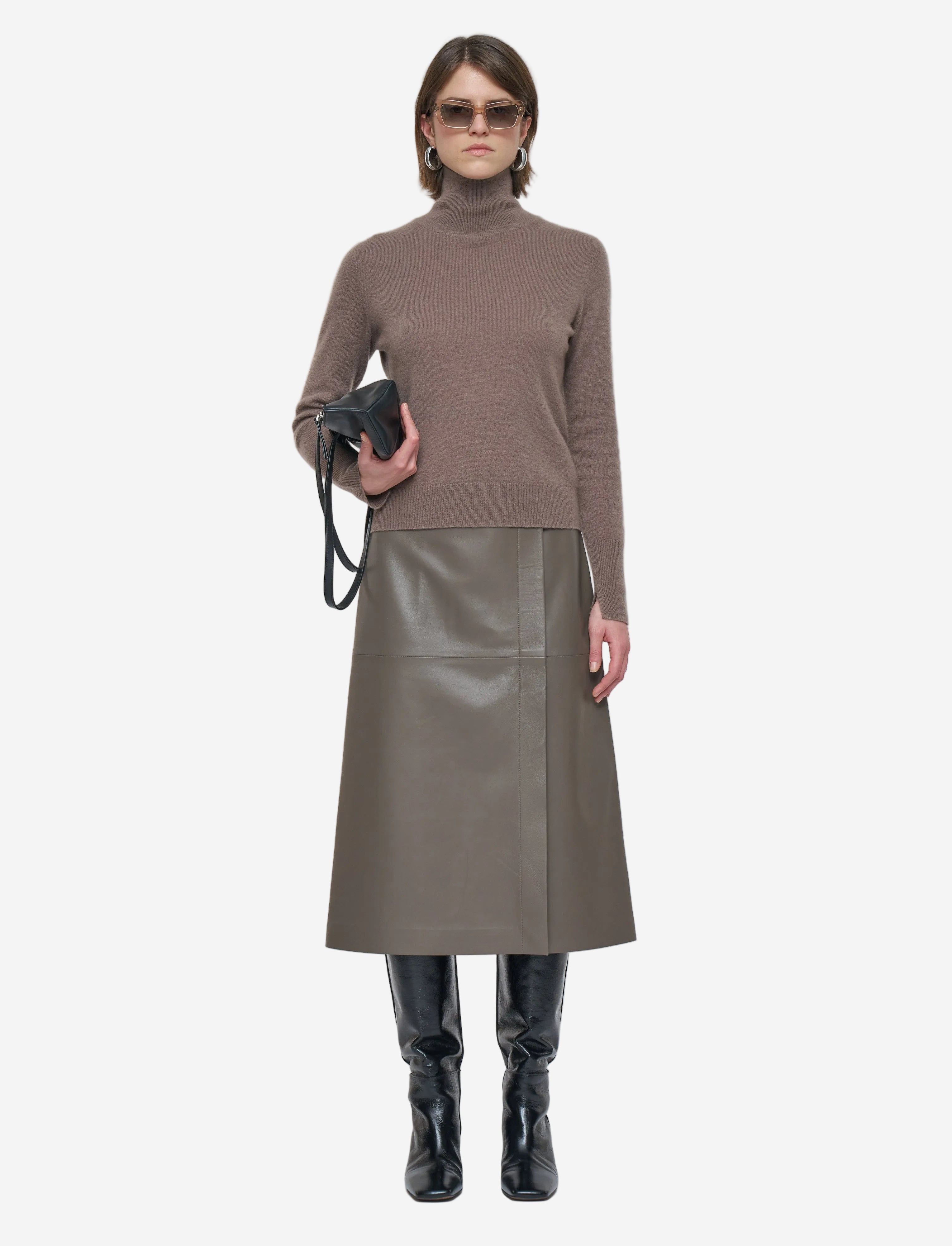 Person wearing a brown turtleneck sweater and leather skirt holding a camera on a white background