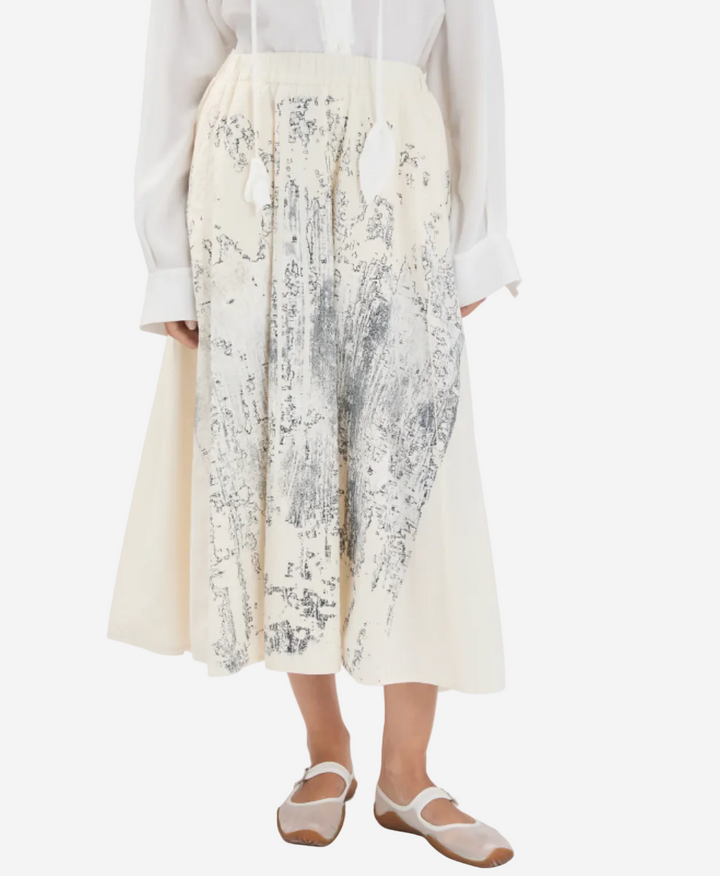Person wearing a white blouse and a long, light-colored skirt with a subtle pattern on a white background