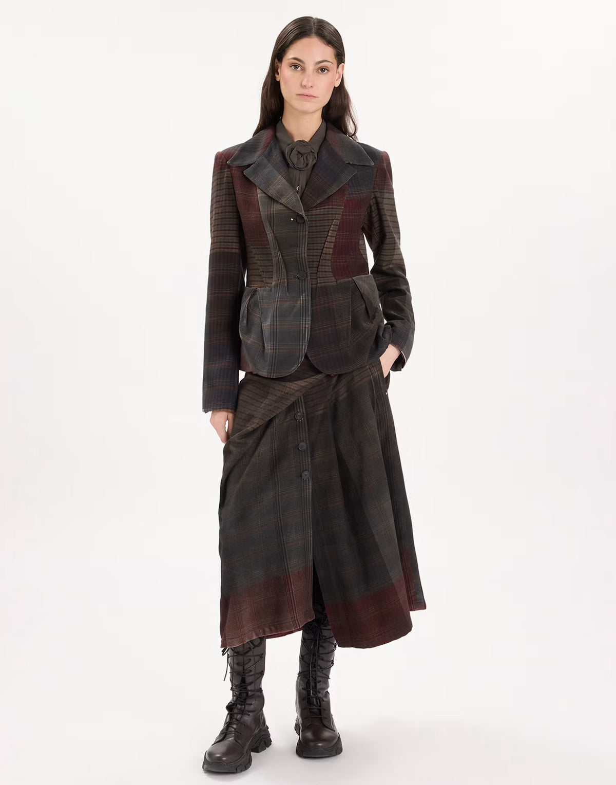 A model stands against a plain white studio background wearing a dark patchwork wool-blend jacket and matching long skirt set with brown leather lace-up boots.