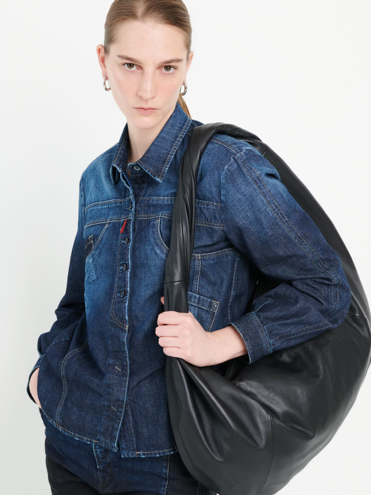 Person wearing a denim jacket holding a black leather bag against a white background