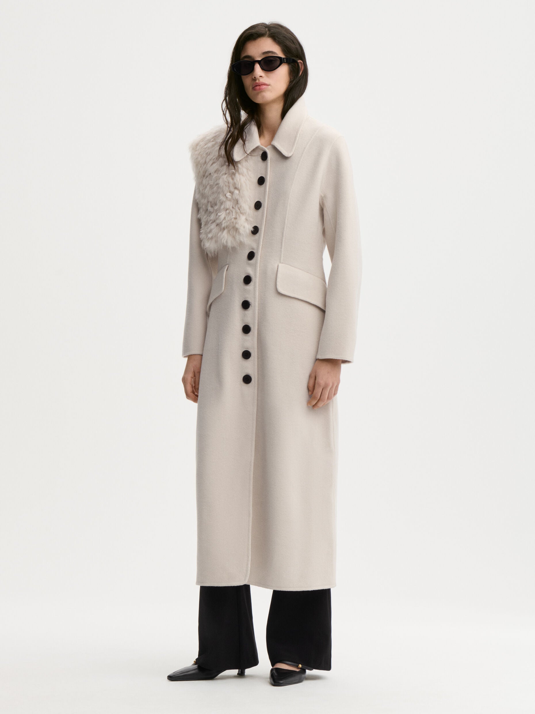 A model wearing a long, light beige wool-blend coat with a textured gray faux-fur collar, black wide-leg trousers, and black pointed-toe flats, photographed against a seamless white studio background.