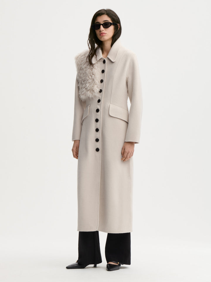 A model wearing a long, light beige wool-blend coat with a textured gray faux-fur collar, black wide-leg trousers, and black pointed-toe flats, photographed against a seamless white studio background.