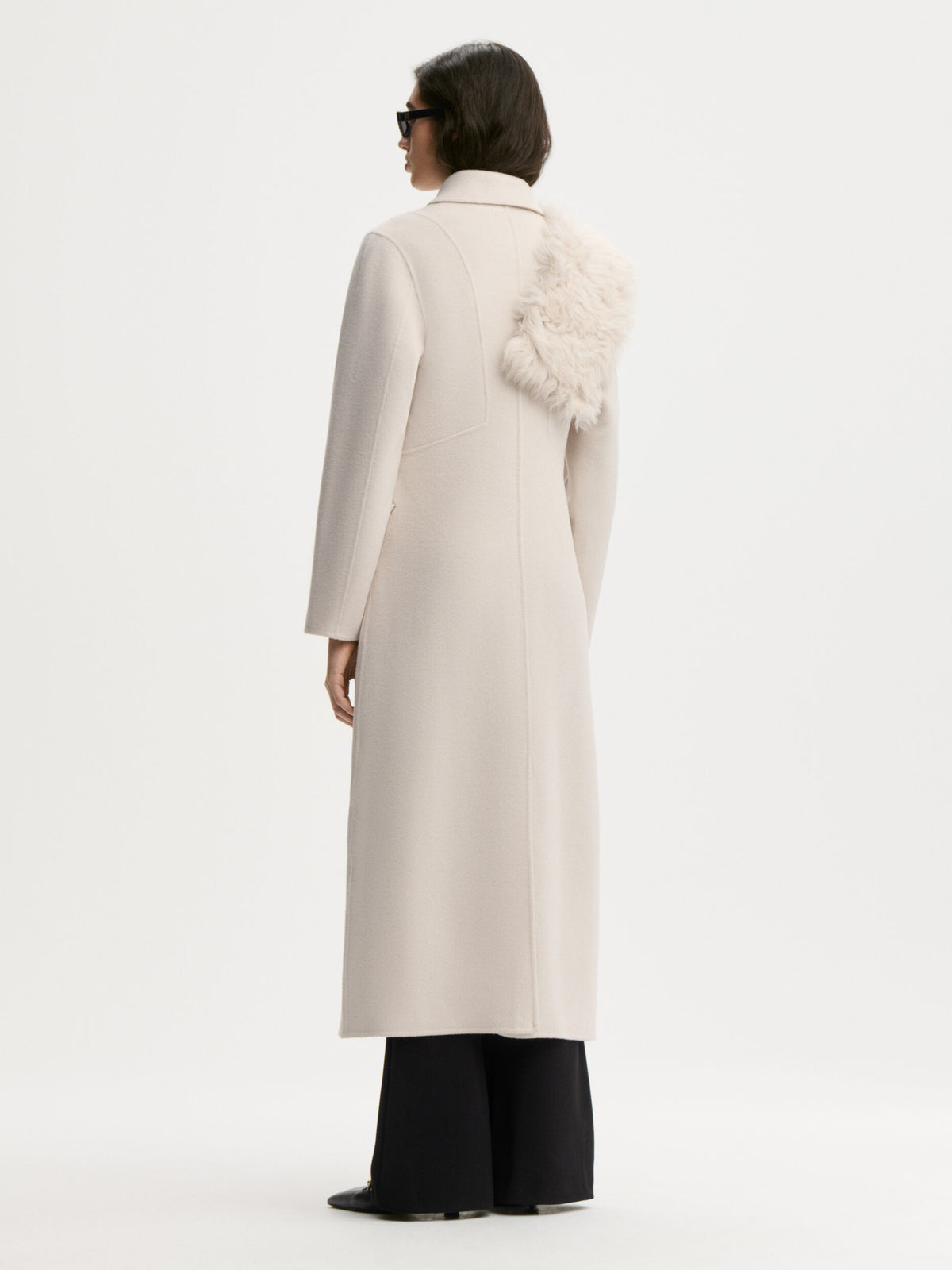 A model shown from the back wearing a long cream-colored coat with a fluffy white faux-fur collar detail, paired with black wide-leg trousers, against a plain white studio background.