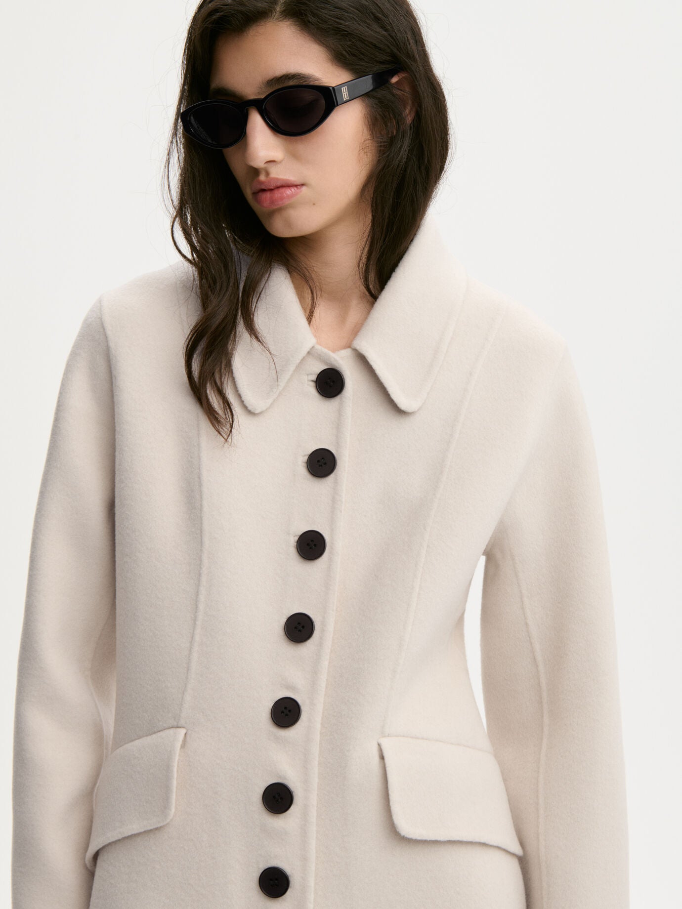 A woman wearing a cream-colored wool coat with large black buttons and black sunglasses, photographed against a clean white studio background.
