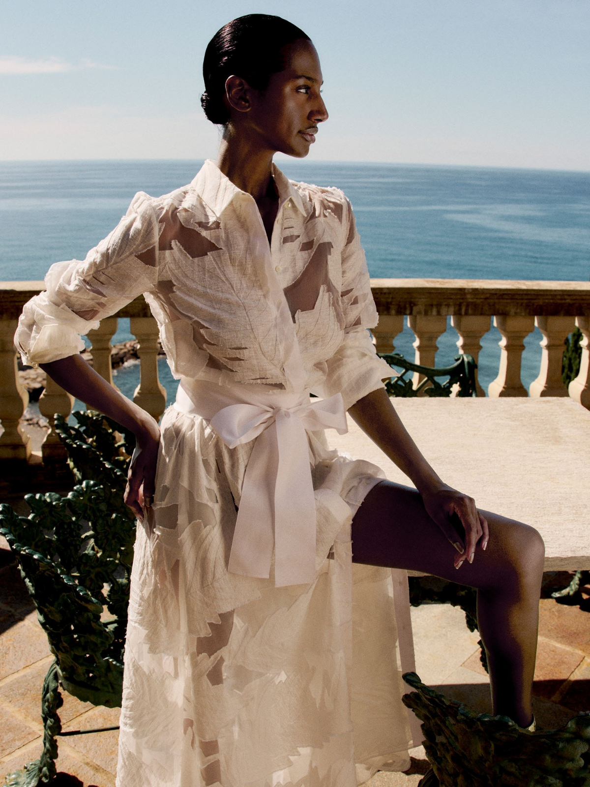Woman in a white dress with brown patterns sitting on a bench by the ocean.