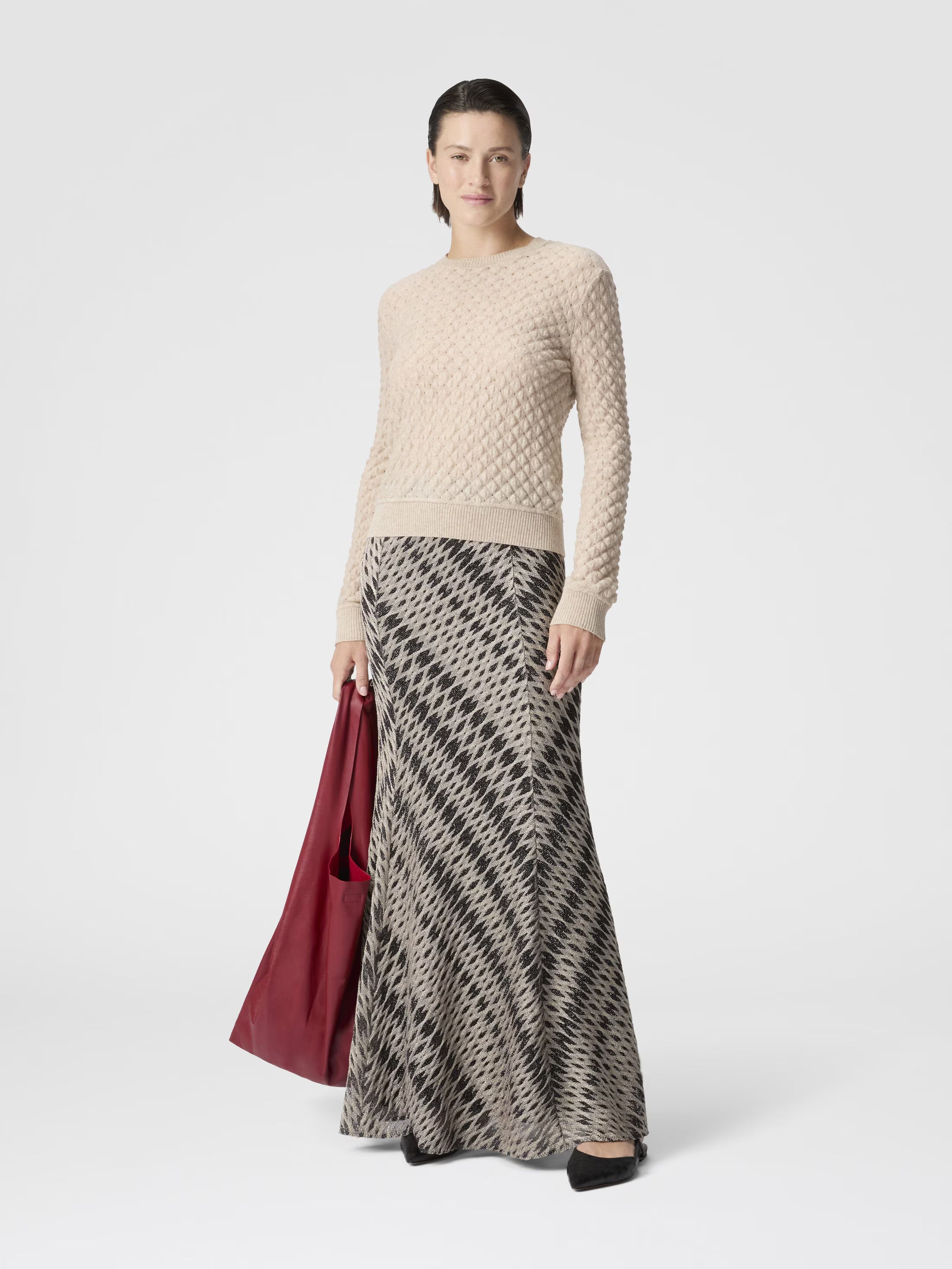 Woman wearing a beige sweater and patterned skirt holding a red bag on a white background