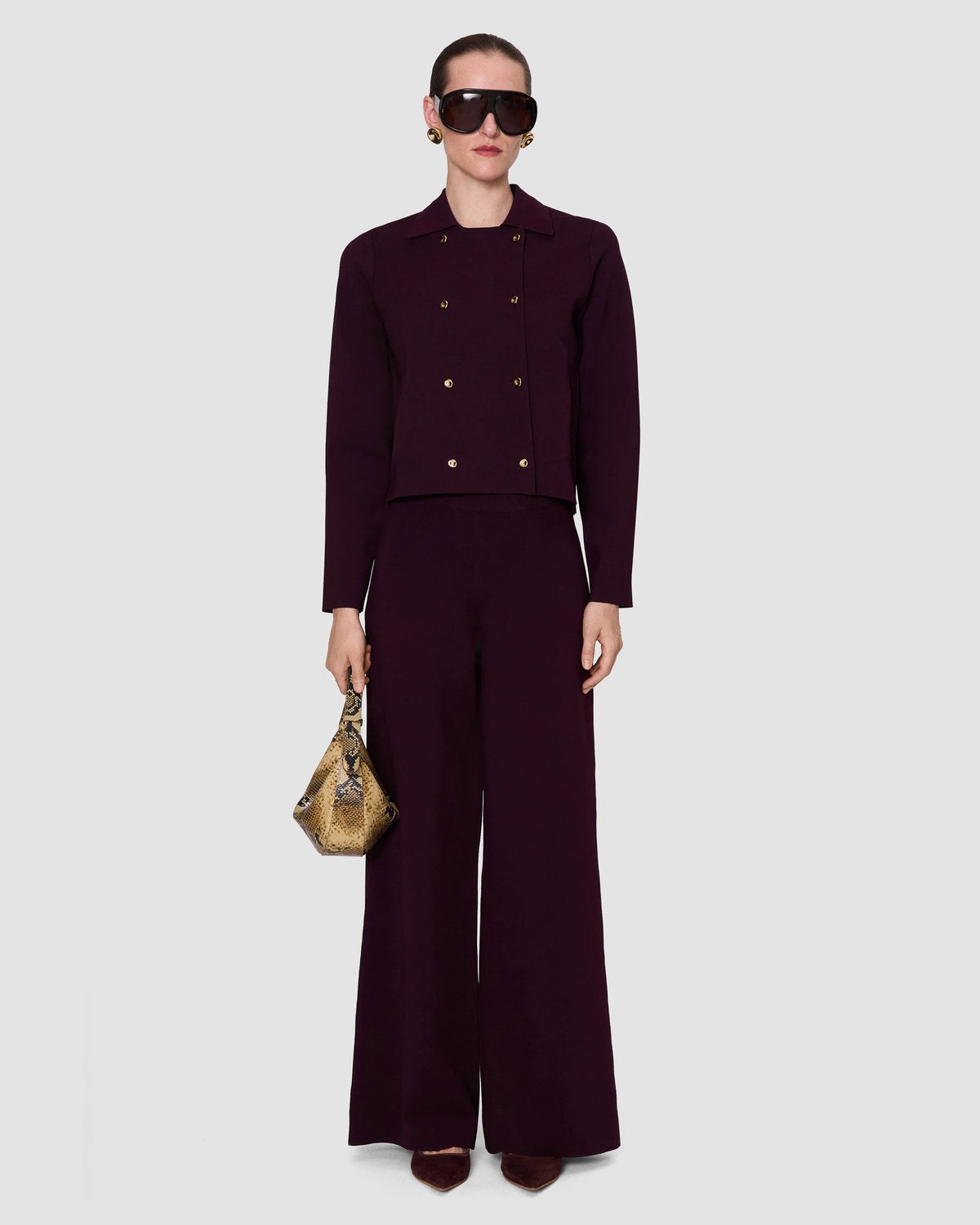 model wearing a deep burgundy double-breasted coat with gold buttons and a structured collar by Joseph, with matching dark purple trousers. 