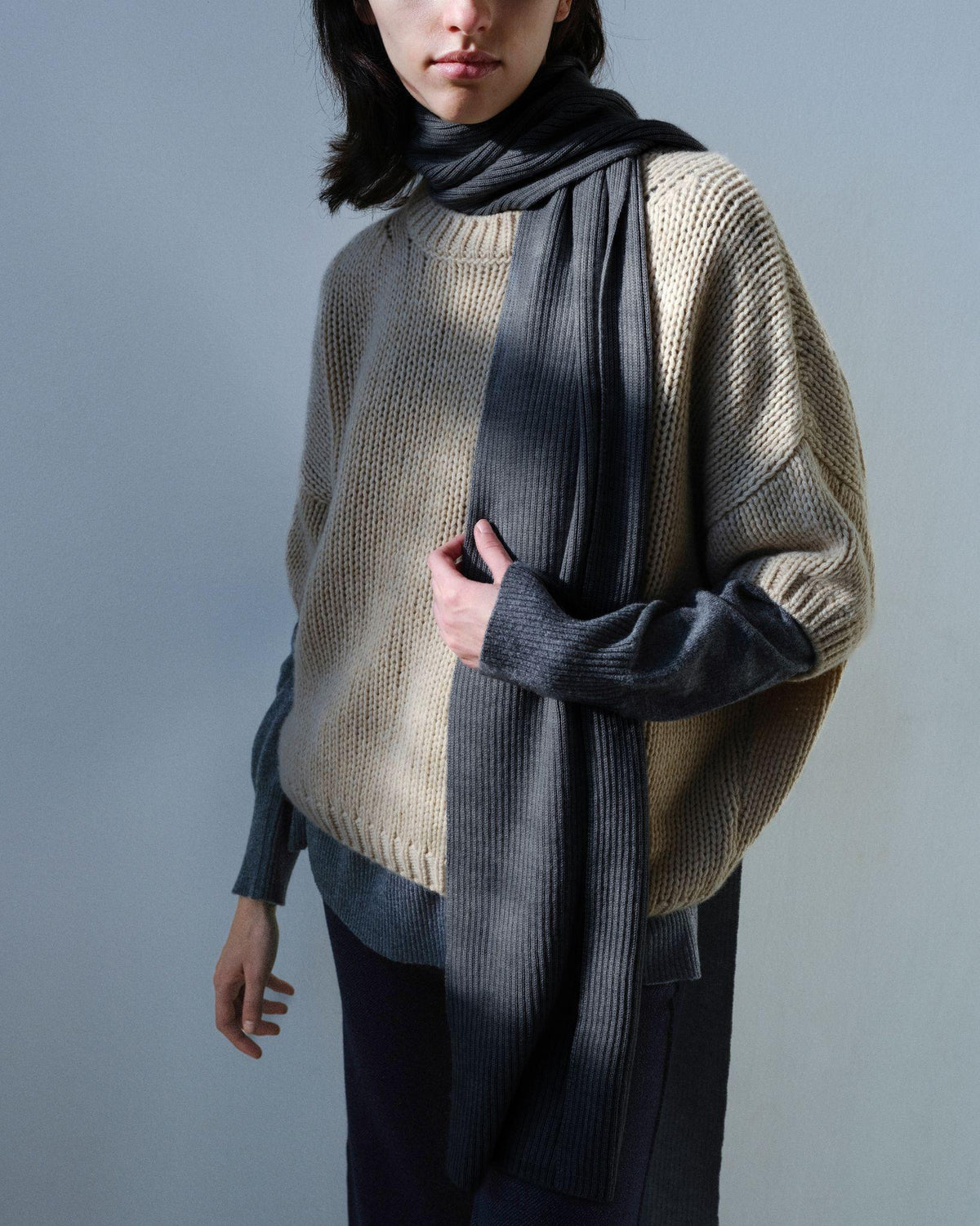A model wearing a chunky knit sweater in beige and dark gray, styled with a matching dark gray ribbed scarf against a solid gray background.