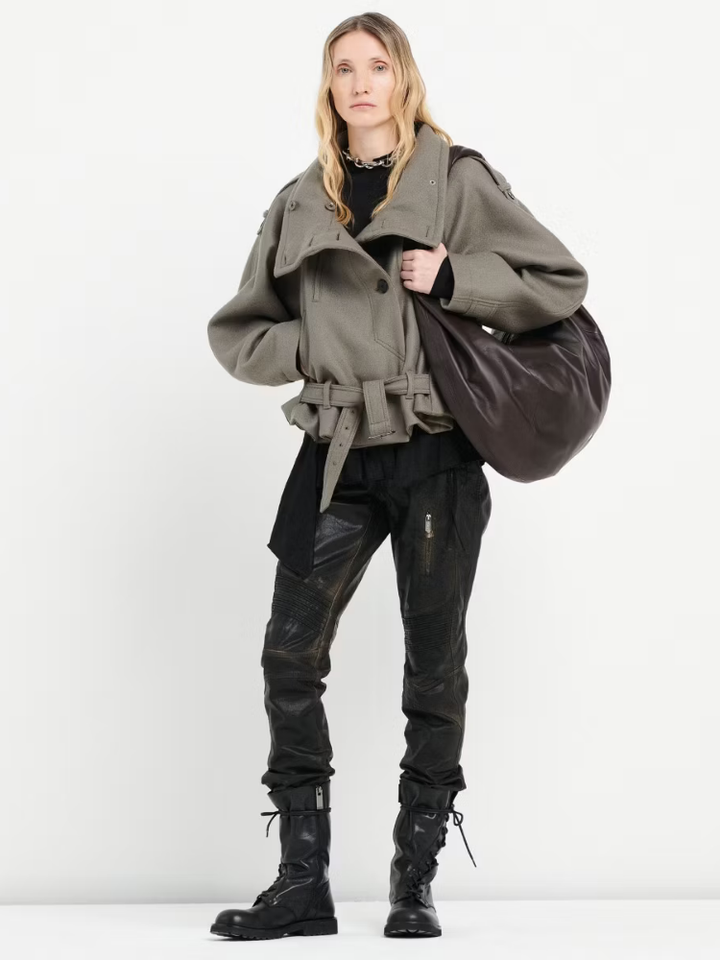 Person wearing a gray coat, black pants, and black boots holding a brown bag on a white background
