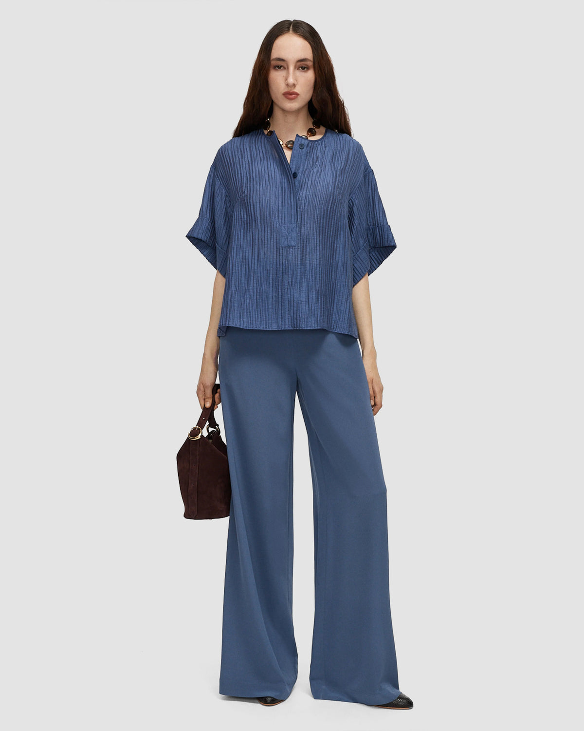 A model wearing a relaxed blue top with a crinkled texture, collarless neckline, partial button front and wide short sleeves with turned-up cuffs by Joseph. 