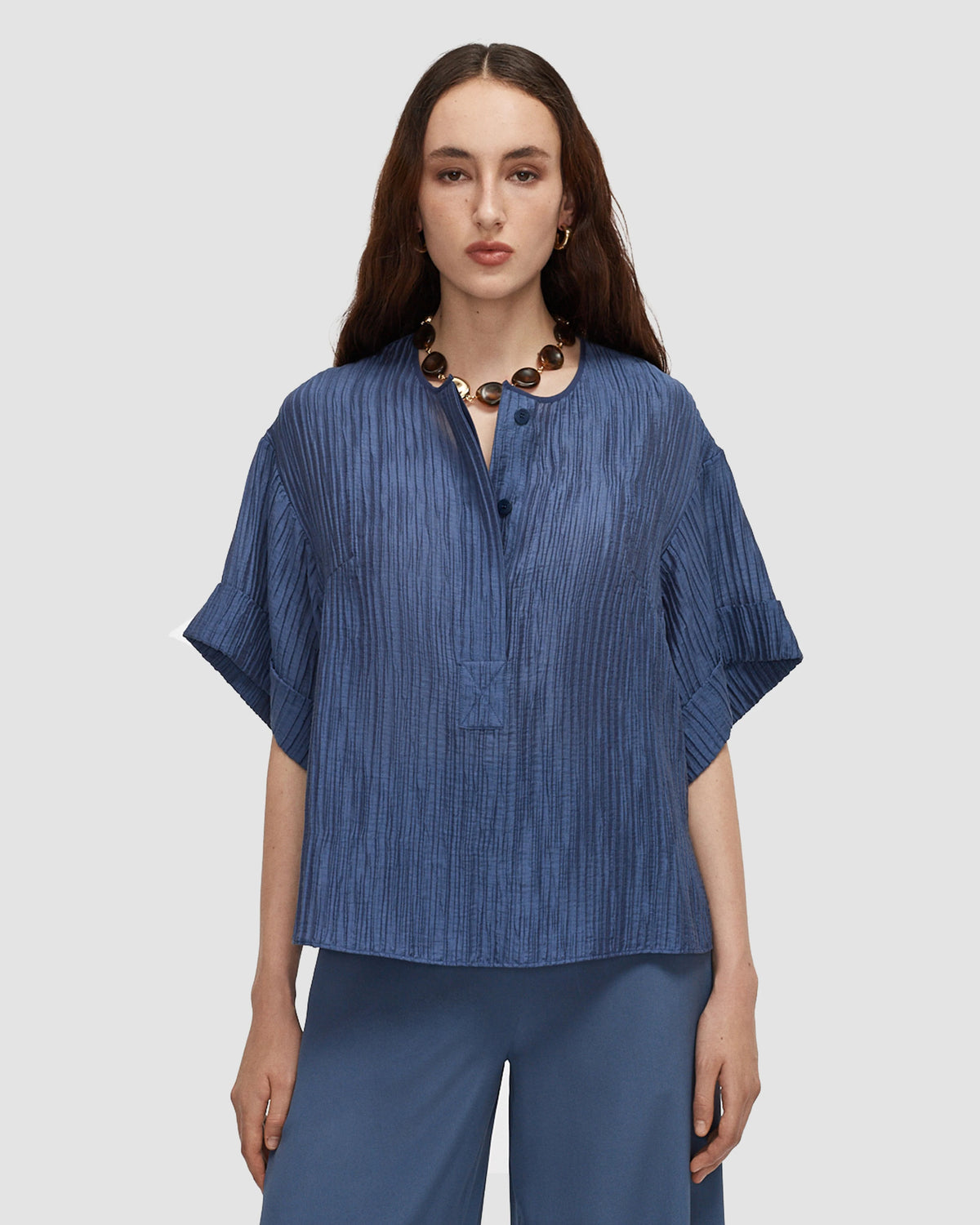 A model wearing a relaxed blue top with a crinkled texture, collarless neckline, partial button front and wide short sleeves with turned-up cuffs from Christensen Copenhagen.