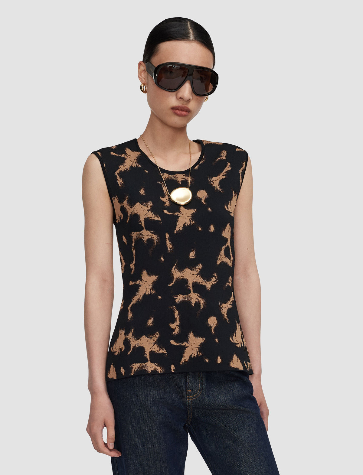 A model wearing a sleeveless black knit top with a tan abstract tortoise pattern, styled with dark sunglasses, a gold pendant necklace, and dark blue jeans against a light grey background.