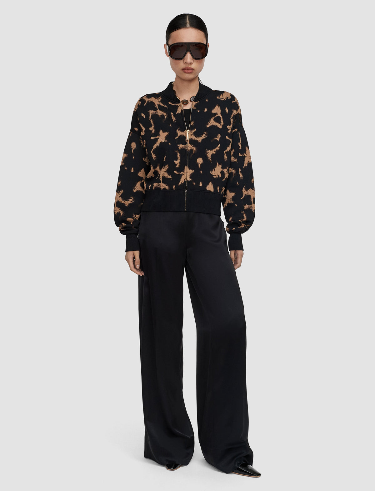 A model wearing a black and tan tortoise patterned bomber jacket from Jospeh with black wide-leg trousers, black sunglasses, and black pointed shoes.
