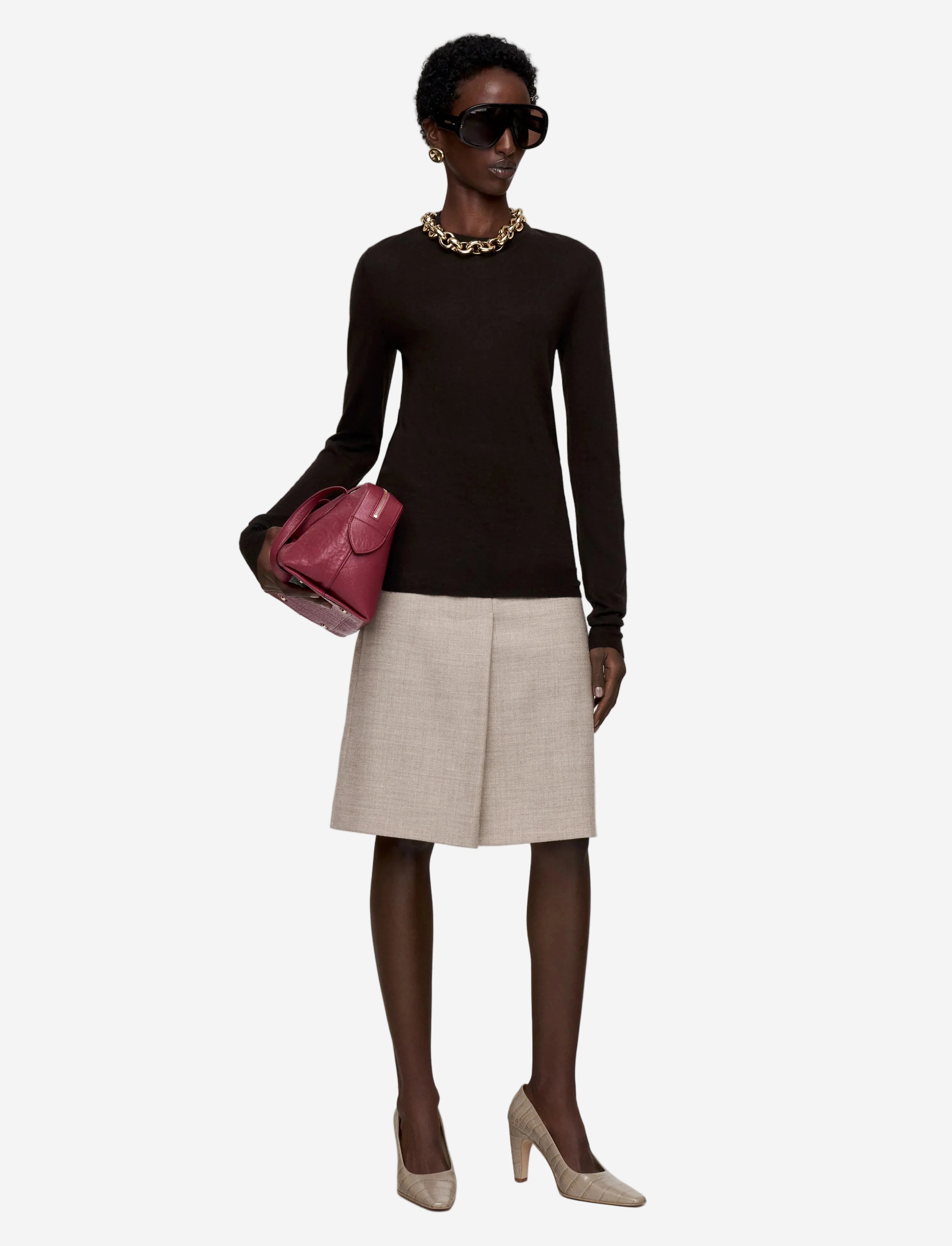 Woman wearing a black long-sleeve top, beige skirt, and holding a pink handbag on a white background