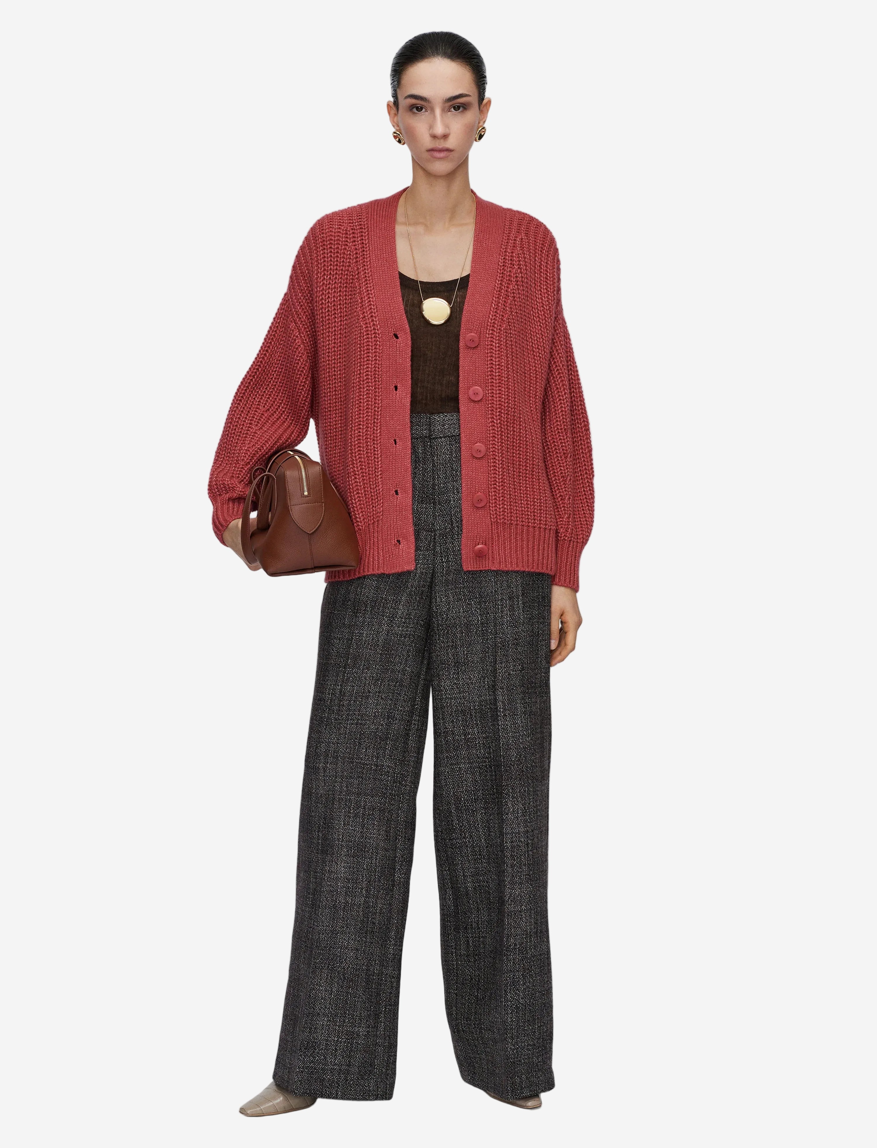 Person wearing a red cardigan, black top, and plaid pants holding a brown bag on a white background