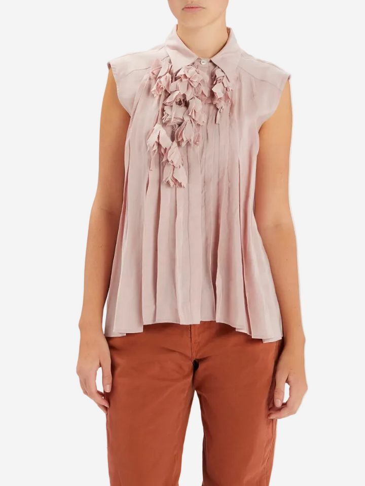 Person wearing a light pink sleeveless blouse with floral details and brown pants on a white background