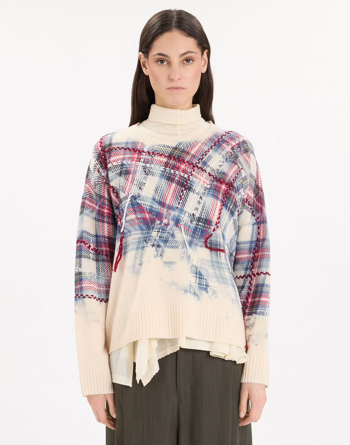 A model wearing a cream-colored knit sweater with a complex multicolor plaid and abstract brushstroke pattern on the front, paired with dark gray trousers, against a plain white studio background.