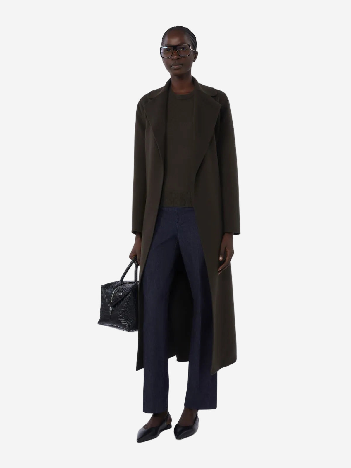 Person wearing a long dark coat and holding a black handbag on a white background