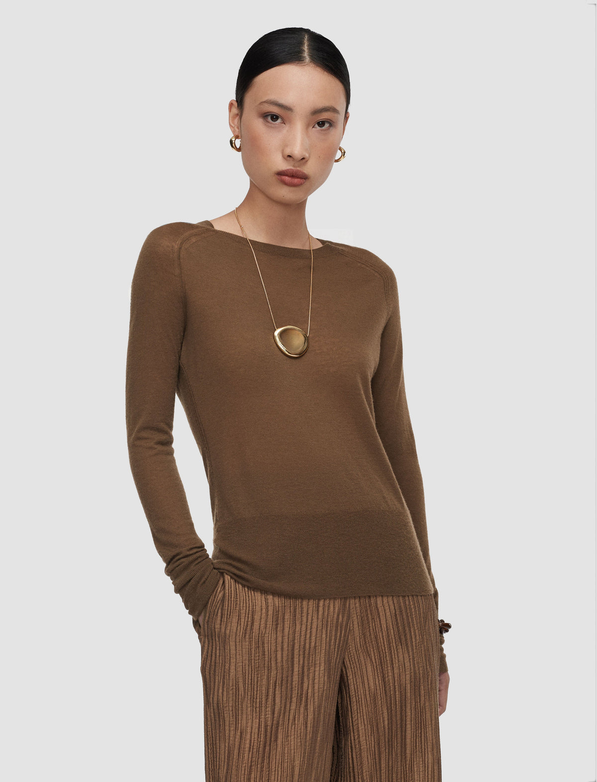 A model wearing a brown long-sleeve knit top from Christensen Copenhagen and pleated brown trousers, accessorized with a gold pendant necklace.
