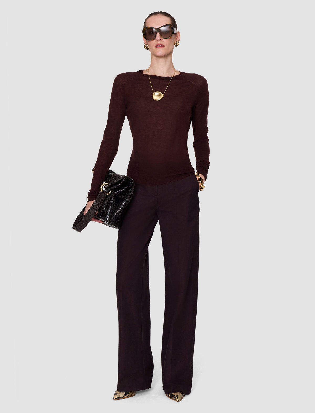 A model wearing a dark burgundy long-sleeve knit top from Joseph and matching wide-leg trousers, styled with gold jewelry, sunglasses, and a black quilted clutch bag. 
