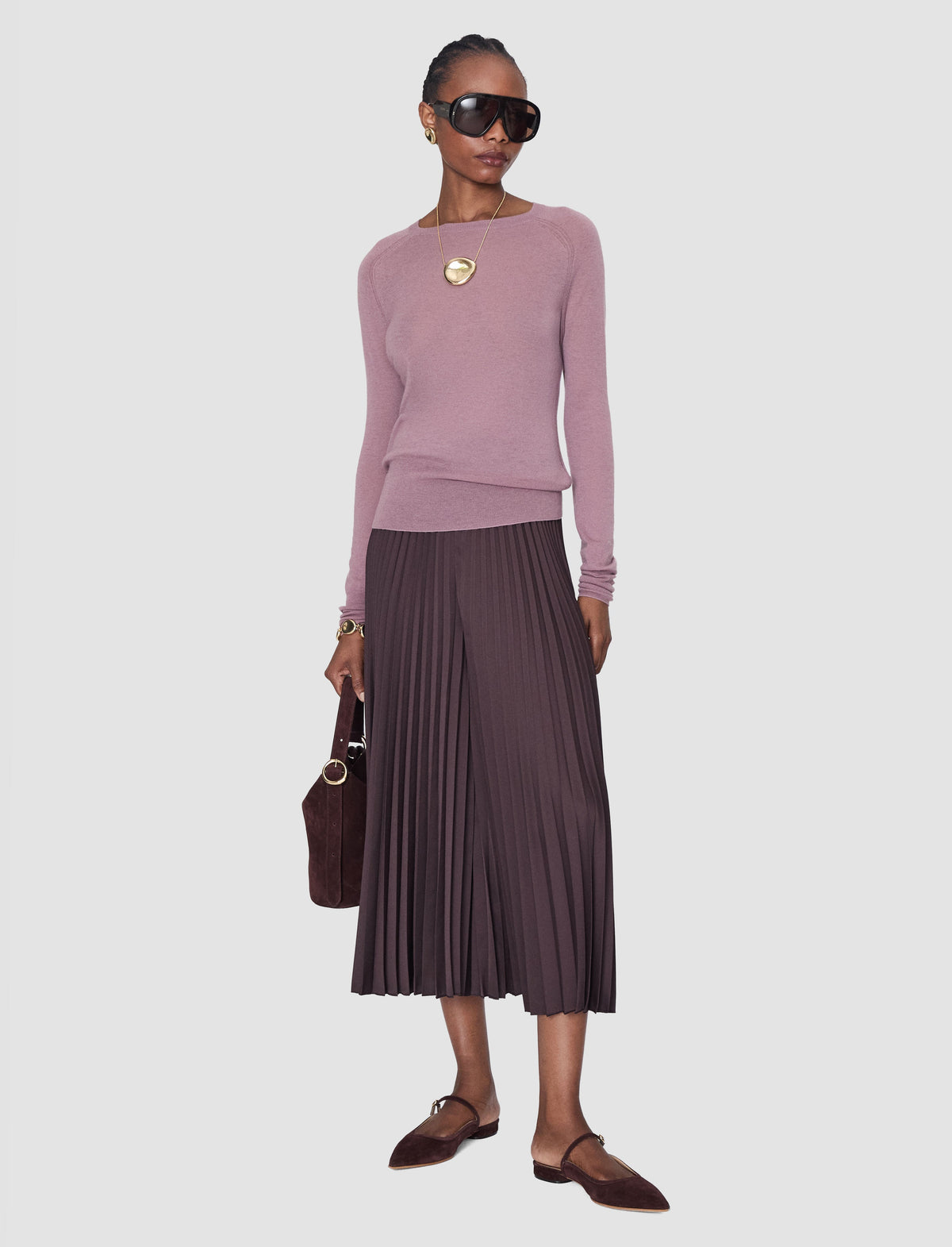 A model is shown wearing a dusty lilac long-sleeve knit jumper by Joseph and a dark pleated midi skirt, styled with brown loafers, a small brown handbag, and a gold pendant necklace.