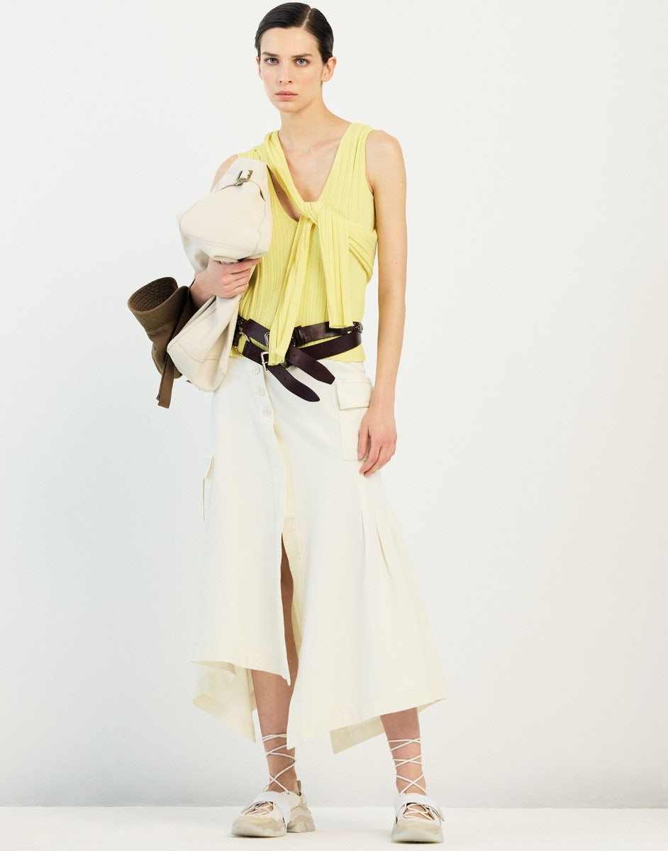 Woman wearing a yellow top and white skirt with a black belt, holding a white bag on a white background.