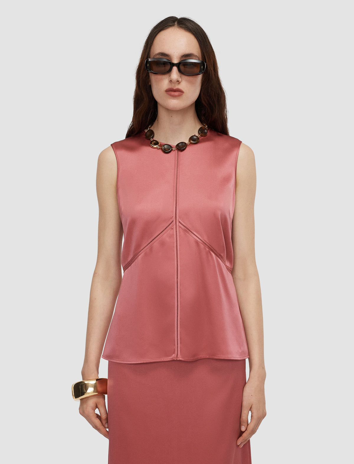 A model wearing a sleeveless pomegranate satin top with a matching skirt, accessorized with a chunky beaded necklace and sunglasses.