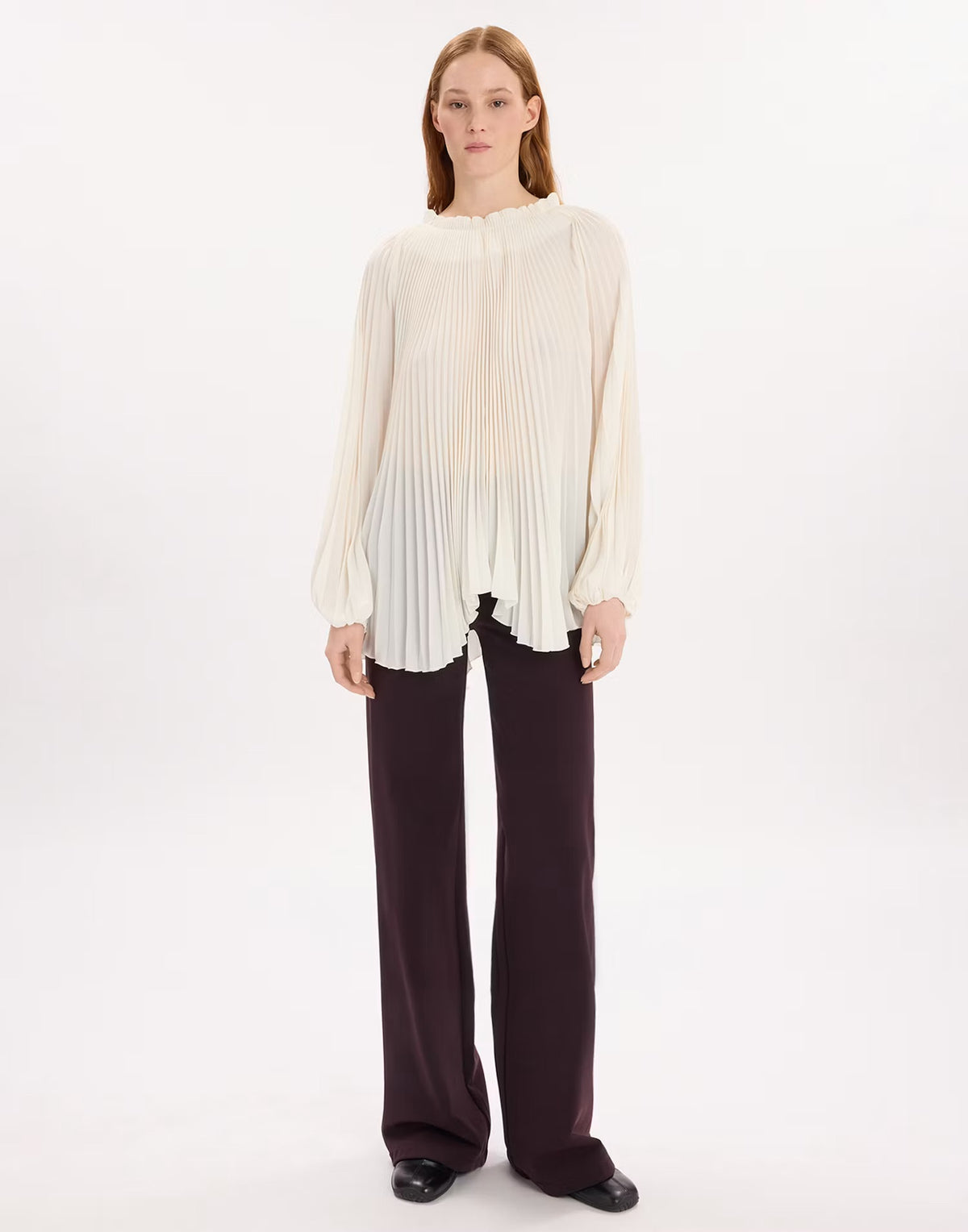 A model stands against a plain white studio background wearing a cream-colored, long-sleeved pleated blouse and dark brown wide-leg trousers.