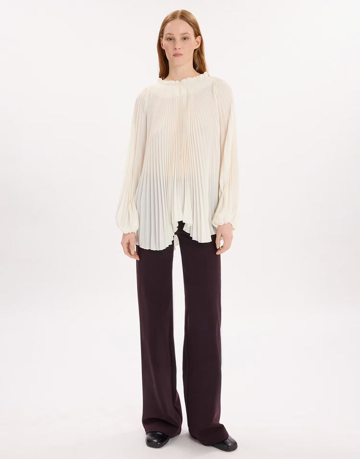 A model stands against a plain white studio background wearing a cream-colored, long-sleeved pleated blouse and dark brown wide-leg trousers.