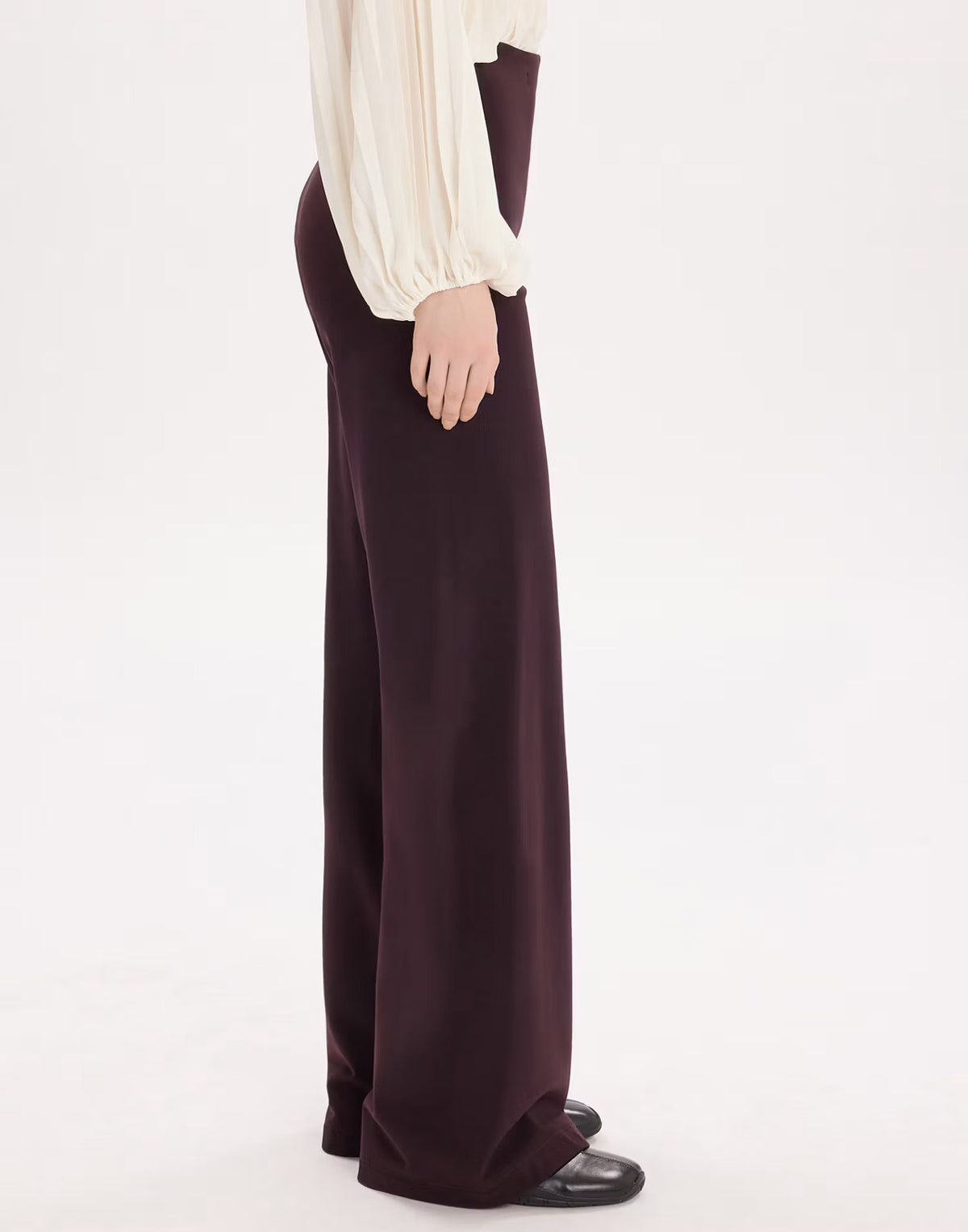 A model is shown from the side wearing dark plum wide-leg trousers, a cream blouse, and black shoes against a plain white studio background.