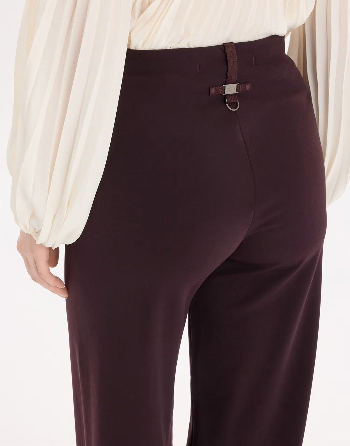 Close-up of a model wearing high-waisted dark plum trousers with a decorative brown leather belt detail and silver buckle at the back waist, paired with a cream blouse.