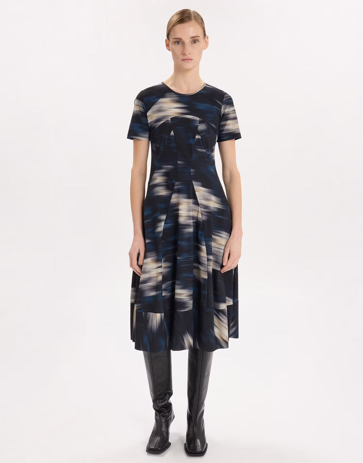 A model stands against a plain white studio background wearing a short-sleeve midi dress with an abstract blue, gray, and cream brushstroke pattern, paired with black knee-high boots.