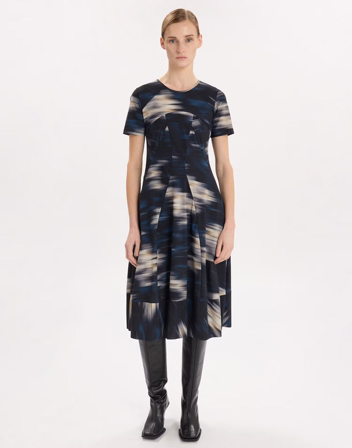 A model stands against a plain white studio background wearing a short-sleeve midi dress with an abstract blue, gray, and cream brushstroke pattern, paired with black knee-high boots.