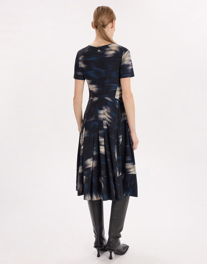 A woman viewed from the back wearing a short-sleeve midi dress with an abstract blue, black, and beige print, paired with black knee-high boots, against a plain white studio background.