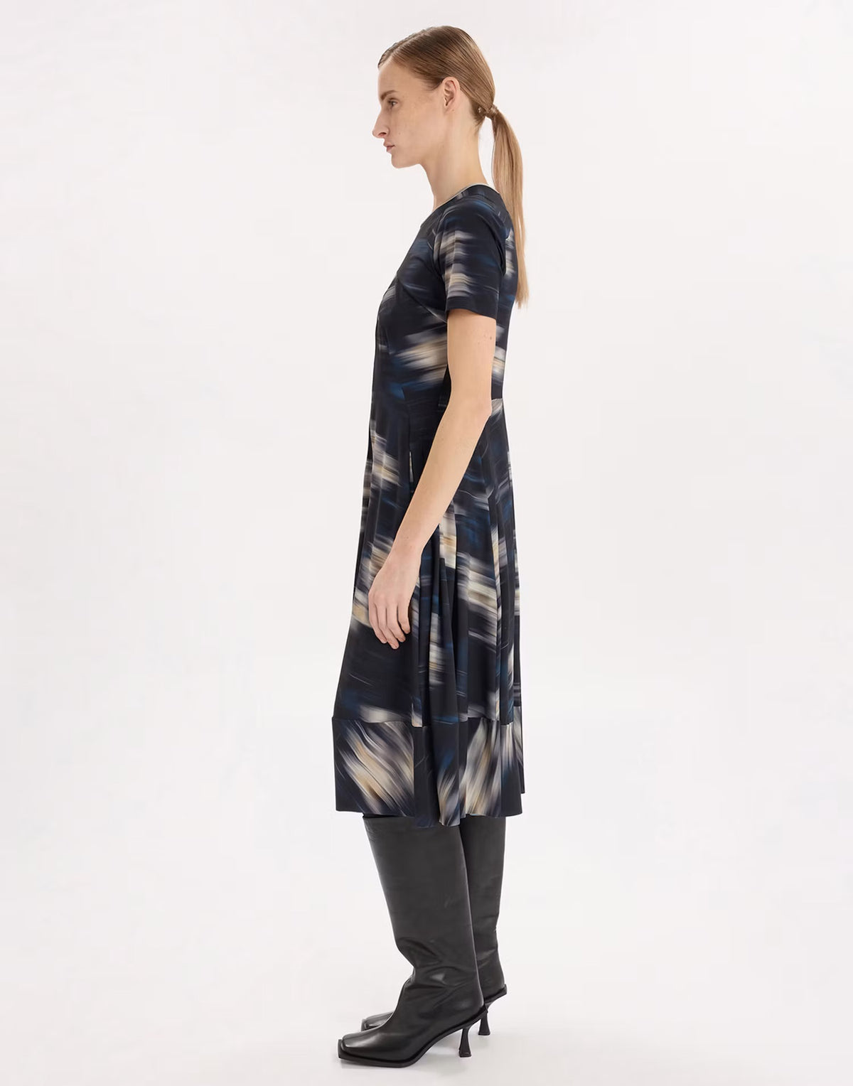 A woman shown in profile wearing a short-sleeve knee-length dress with an abstract navy, beige, and gray print, paired with black pants and black heeled boots, against a plain white studio background.