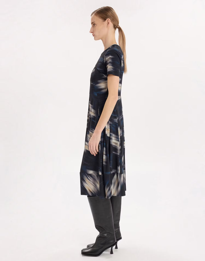 A woman shown in profile wearing a short-sleeve knee-length dress with an abstract navy, beige, and gray print, paired with black pants and black heeled boots, against a plain white studio background.