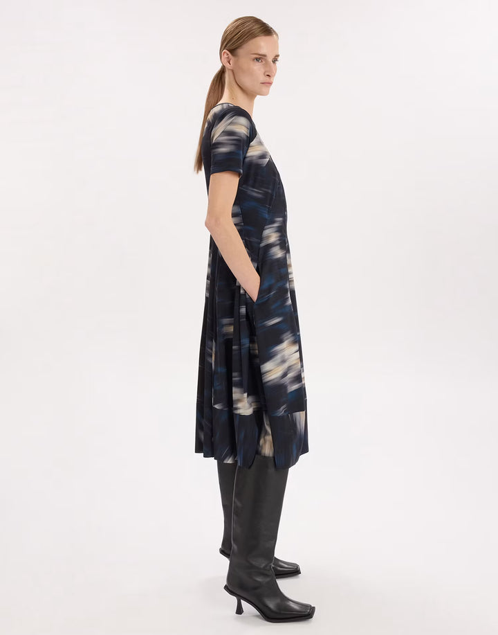 A woman modeling a short-sleeve, knee-length dress with an abstract blue, navy, and beige pattern, paired with black knee-high boots, shown from a side profile against a plain white studio background.