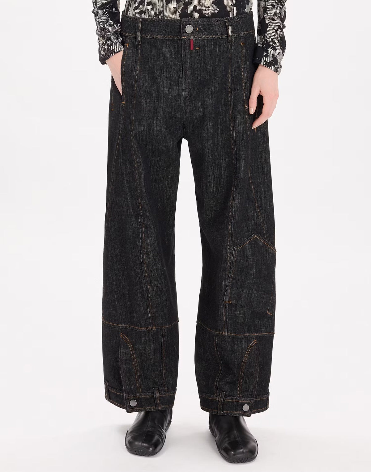 A person wearing very wide-leg dark indigo denim jeans with contrast orange-brown stitching, paired with black boots and a patterned long-sleeve top, shown from the waist down on a white studio background.