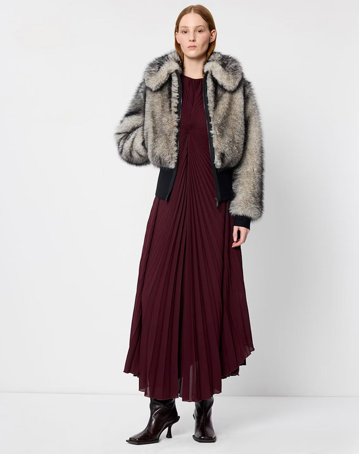 A model stands against a light gray studio background wearing a dark burgundy pleated maxi dress with a large gray and black faux fur collar jacket, paired with dark brown ankle boots.