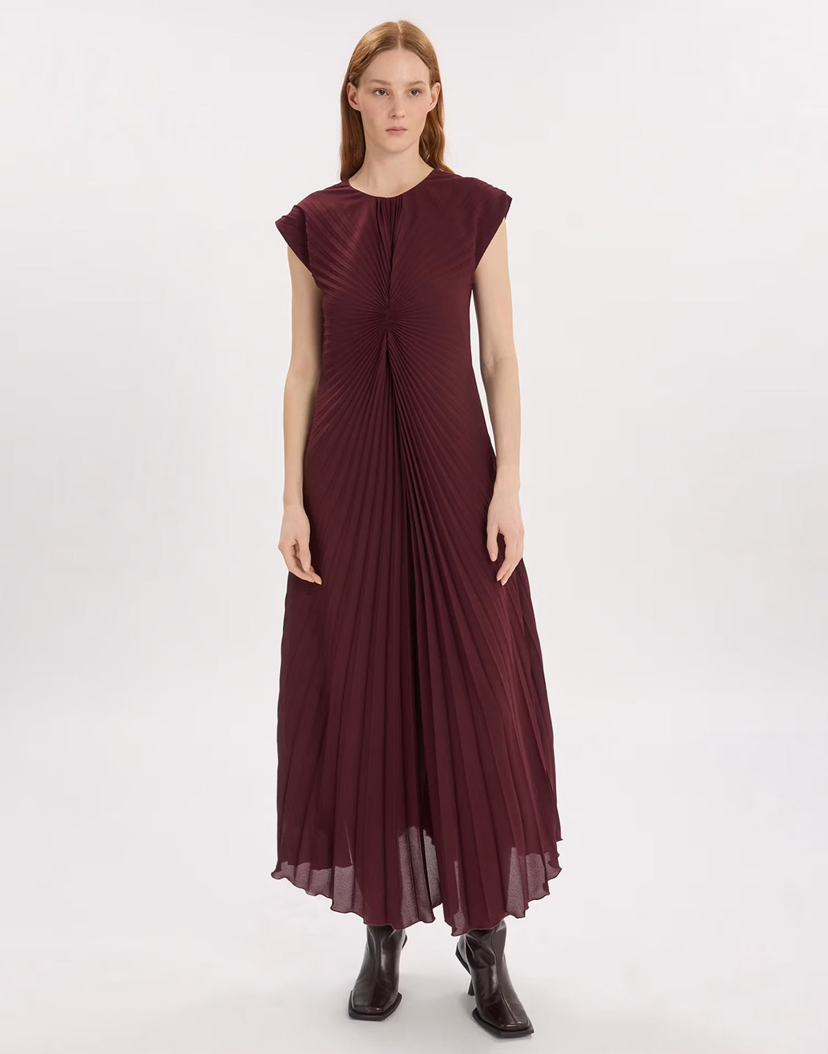 A woman modeling a sleeveless, floor-length pleated dress in deep burgundy, paired with dark brown ankle boots, against a plain white studio background.