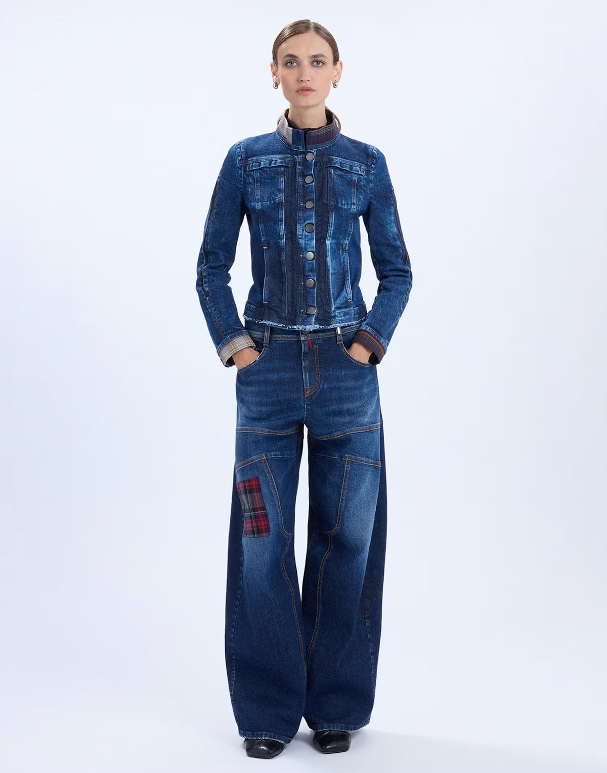 A model stands against a light gray studio background wearing a dark blue denim jacket and matching wide-leg denim pants with a red plaid patch on the left leg.