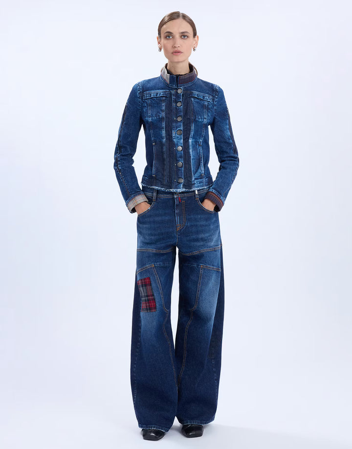 A model stands against a light gray studio background wearing a dark blue denim jacket and matching wide-leg denim pants with a red plaid patch on the left leg.