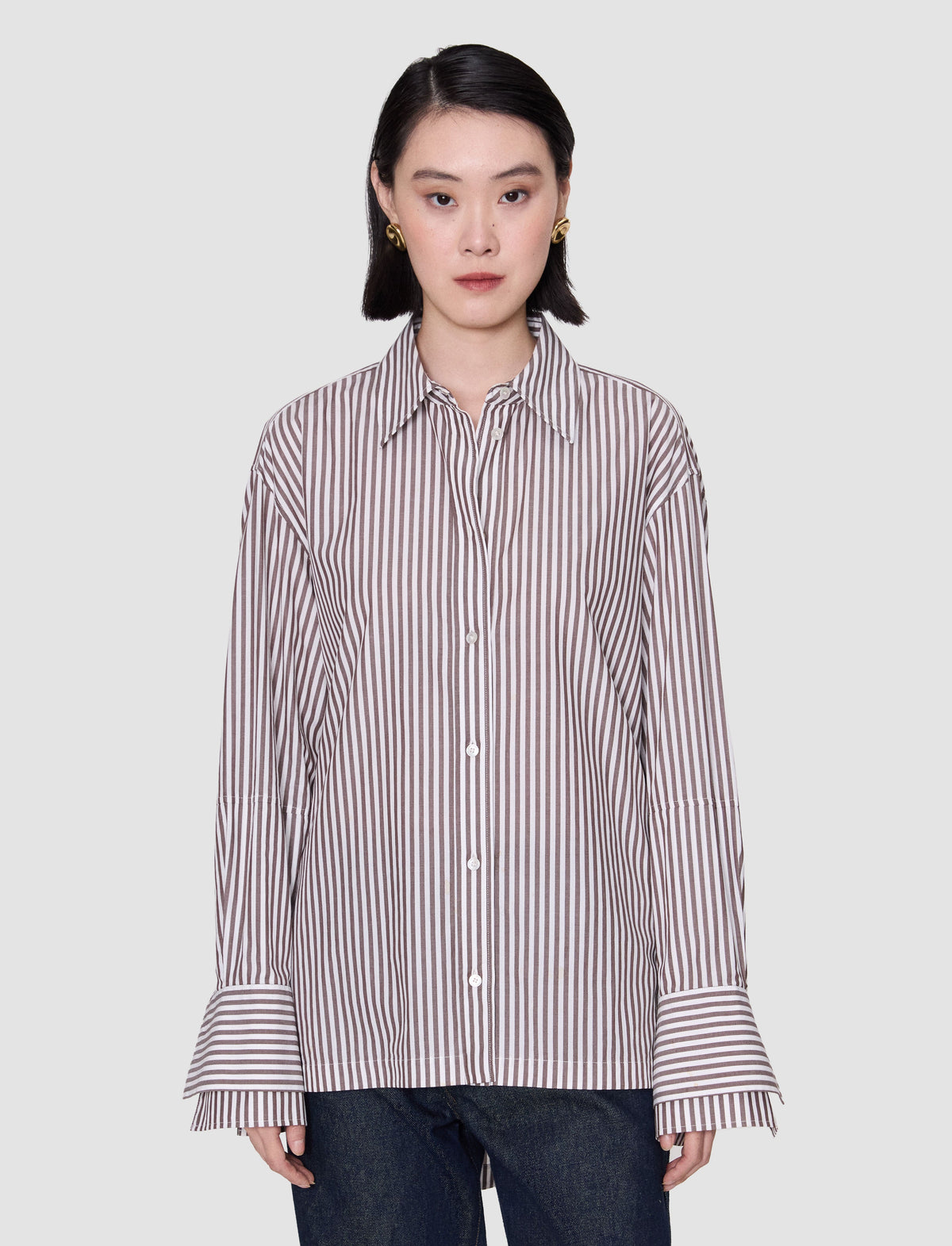 A model wearing an oversized grey and white striped shirt with a pointed collar, button front, dropped shoulders, wide cuffs and a longer back hem. 
